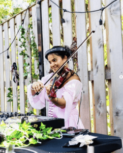 Female DJ playing violin at outdoor event with DJ equipment and greenery.